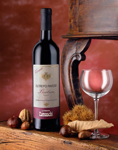 Foto del Vino Rosso Barbera Comaschi con scenografia: Bicchiere,Frutta Secca,Sfondo Rosso e Tavolinof
