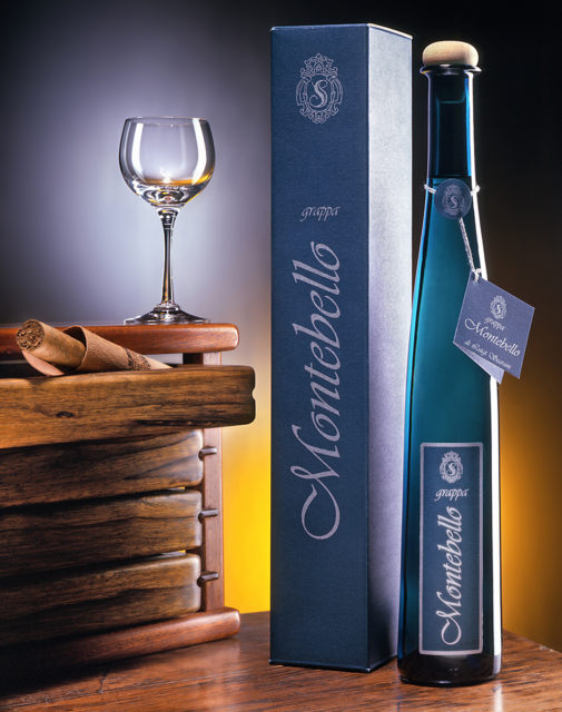 Foto still life della Grappa Ca'Montebello con bicchiere,mobiletto,sigaro, su fondo grigio-arancio