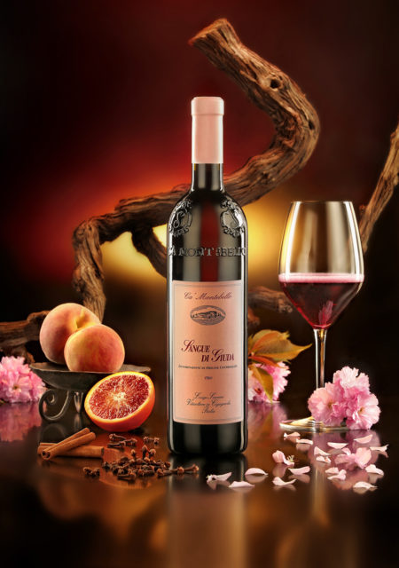 Foto still life del vino rosso Sangue di Giuda con bicchiere, fiori,spezie,frutta, su fondo rosso e tralcio.